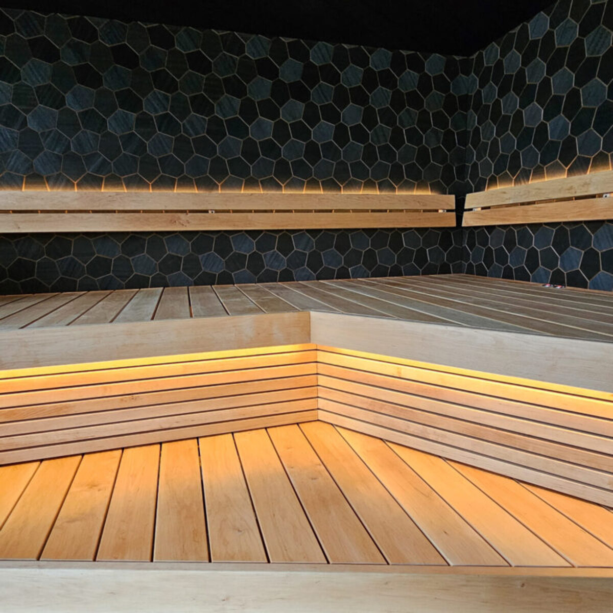 Sauna corner with dark hexagon wall paneling and a backlit light wood bench